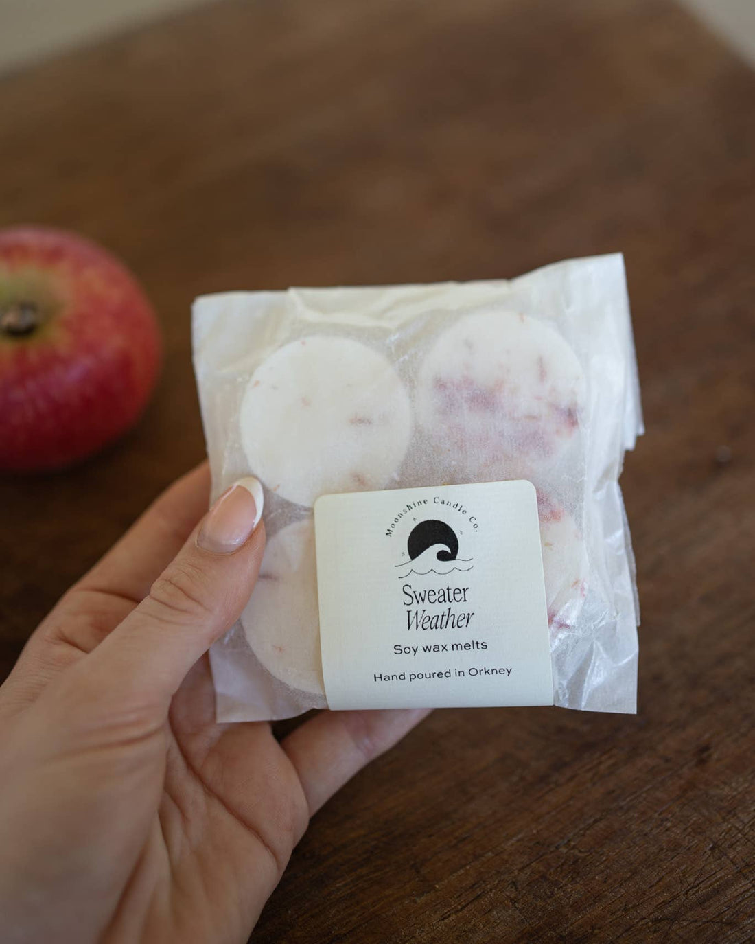 Hand holding a package of Sweater Weather soy wax melts on a wooden surface with an apple in the background.