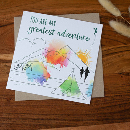 Handmade square greeting card with &quot;You Are My Greatest Adventure&quot; text, featuring a couple running up a mountain in line art with watercolour splashes. Eco-conscious and packaged with a Kraft envelope.