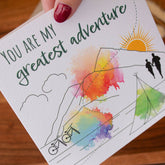 Handmade square greeting card with "You Are My Greatest Adventure" text, featuring a couple running up a mountain in line art with watercolour splashes. Eco-conscious and packaged with a Kraft envelope.