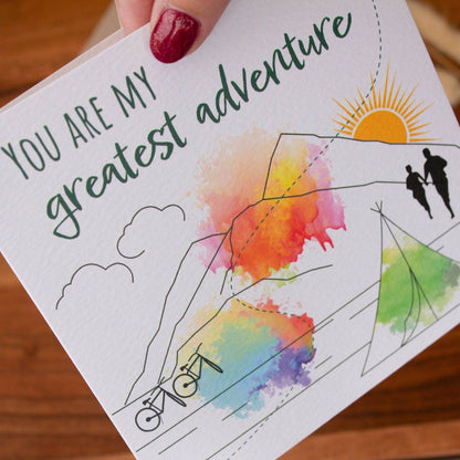 Handmade square greeting card with &quot;You Are My Greatest Adventure&quot; text, featuring a couple running up a mountain in line art with watercolour splashes. Eco-conscious and packaged with a Kraft envelope.