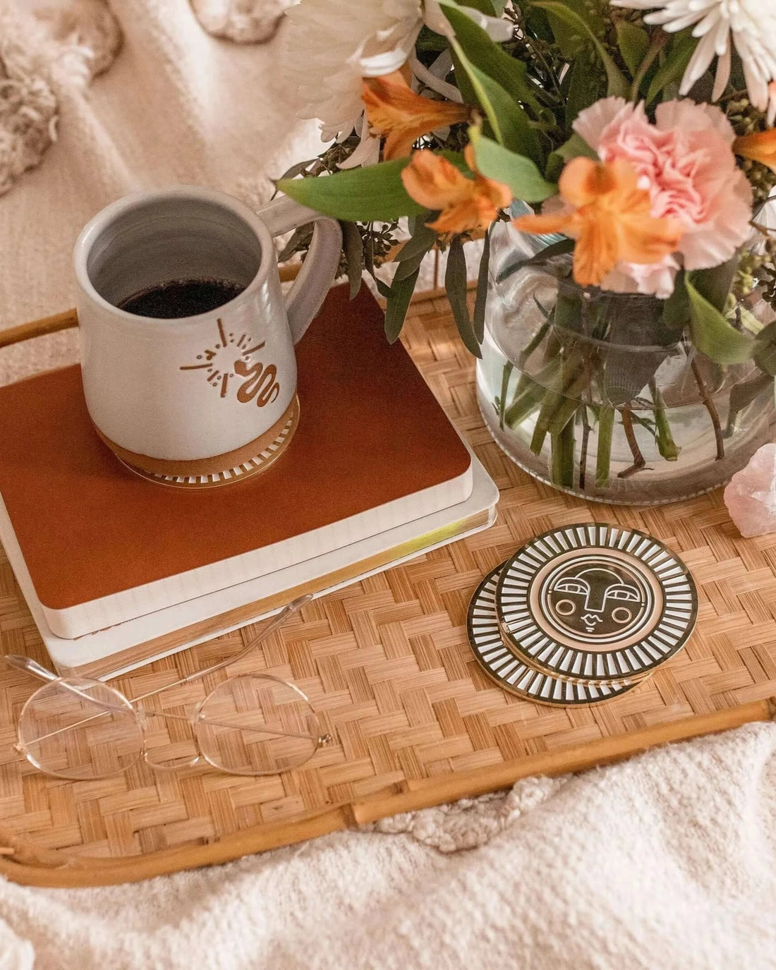 Set of two handmade round warm beige coasters with gold-coloured metal and enamel, showcasing the Sol face design. On a tray in lifestyle shot