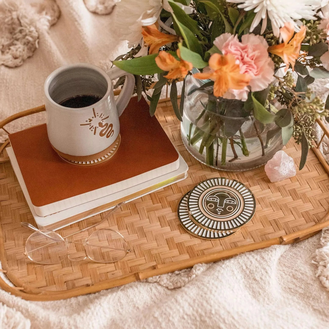 Set of two handmade round warm beige coasters with gold-coloured metal and enamel, showcasing the Sol face design. On a tray in lifestyle shot