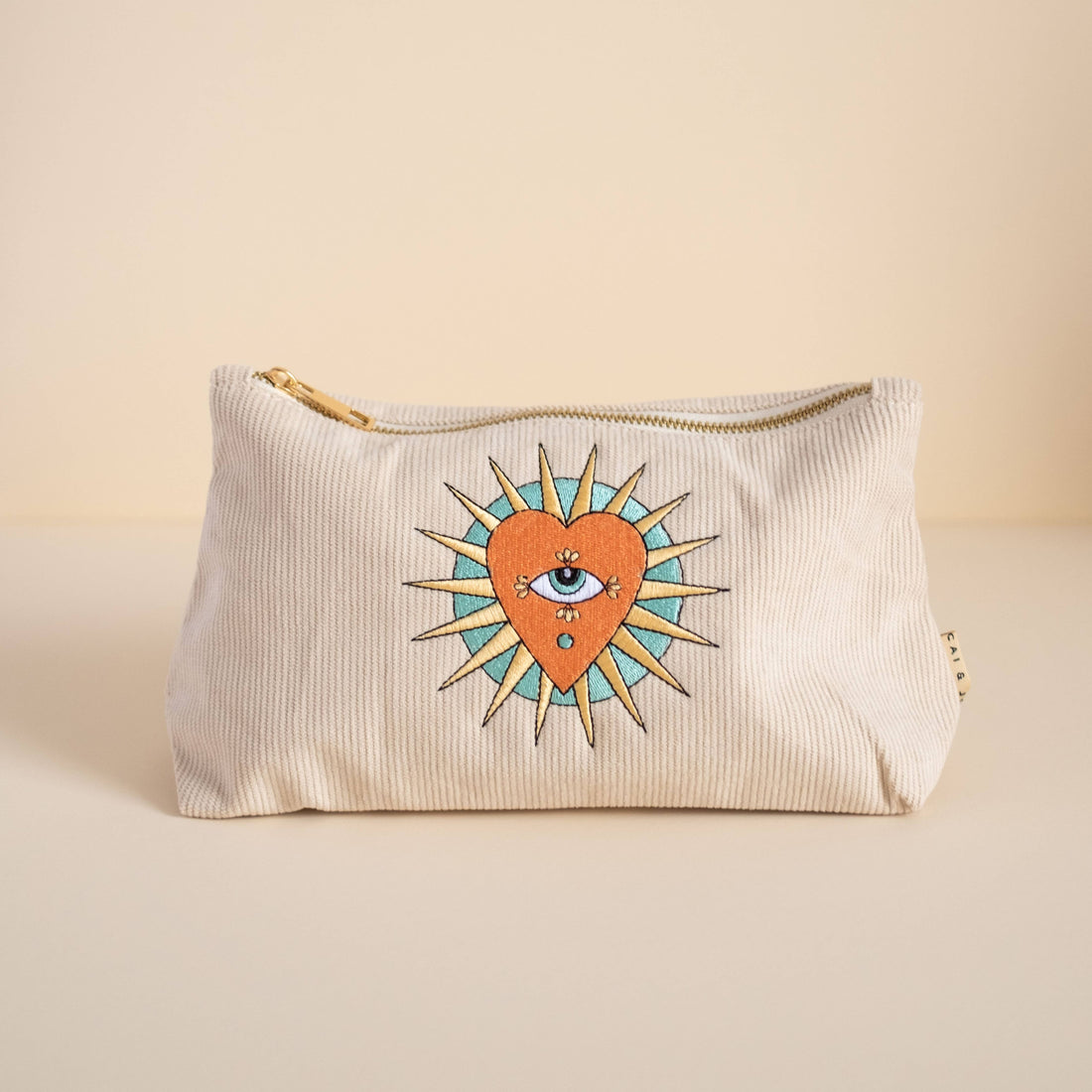 Stone-coloured medium corduroy pouch with a zip closure, featuring embroidered designs of a heart, eye, and sun. Fully lined for storing makeup, toiletries, or travel accessories.