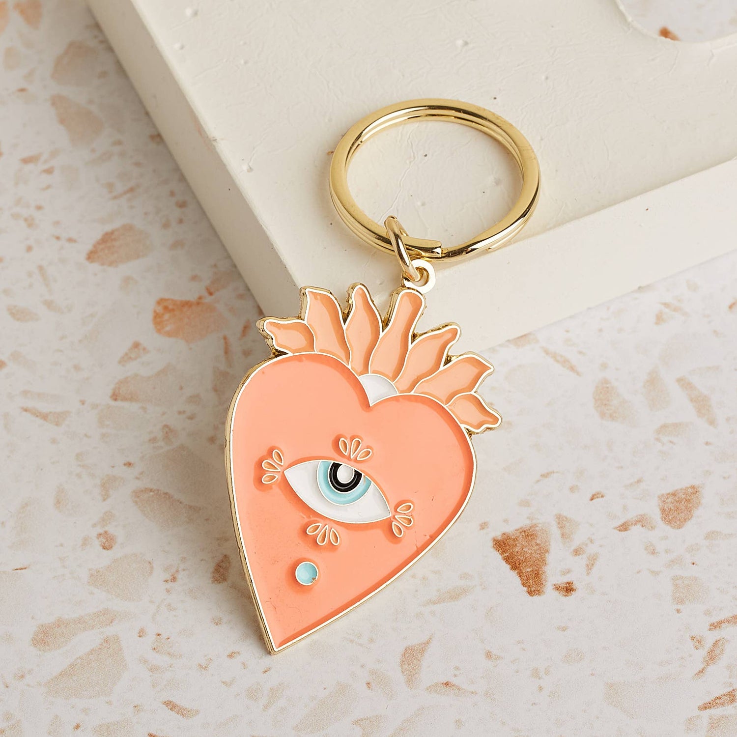 Soft enamel keychain with a pale orange heart design featuring an eye in the centre, accented with gold highlights. Includes a matching gold ring.