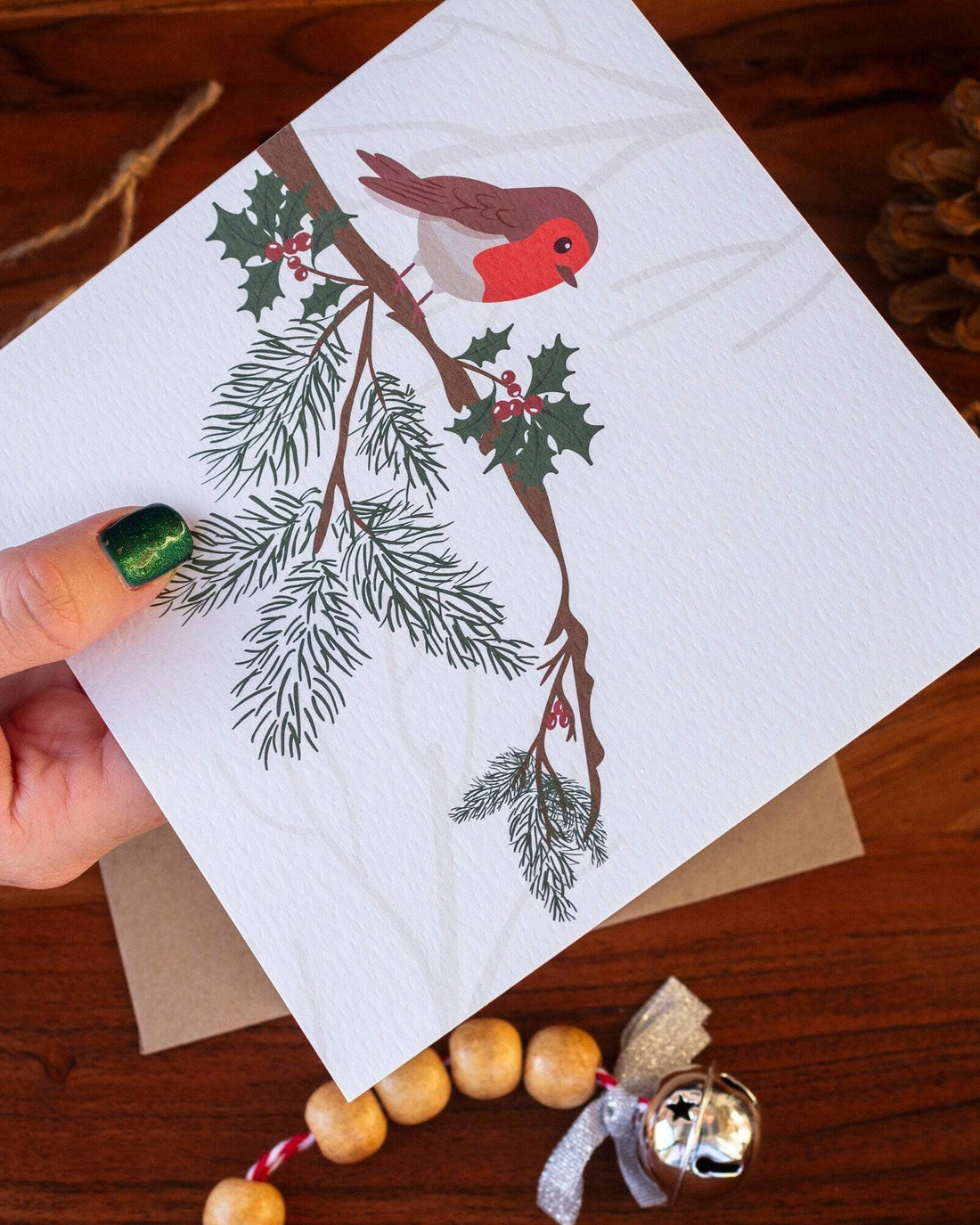 Handmade square Christmas card featuring a robin perched on a holly branch, printed on sustainably sourced textured paper with a biodegradable cello wrap and Kraft envelope.