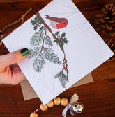 Handmade square Christmas card featuring a robin perched on a holly branch, printed on sustainably sourced textured paper with a biodegradable cello wrap and Kraft envelope.