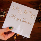 Square Christmas card with white snowy trees, hanging gold and white baubles, and "Merry Christmas" written in gold. Made from recyclable luxury textured paper with eco-friendly packaging.